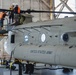 Maintenance personnel are working on a United States Army CH-47 Chinook helicopter inside a large hangar facility. Three workers wearing orange safety vests and hard hats are positioned on an elevated work platform accessing the rotor head assembly at the top of the aircraft. A fourth worker in an orange vest is visible on the hangar floor in the background.
