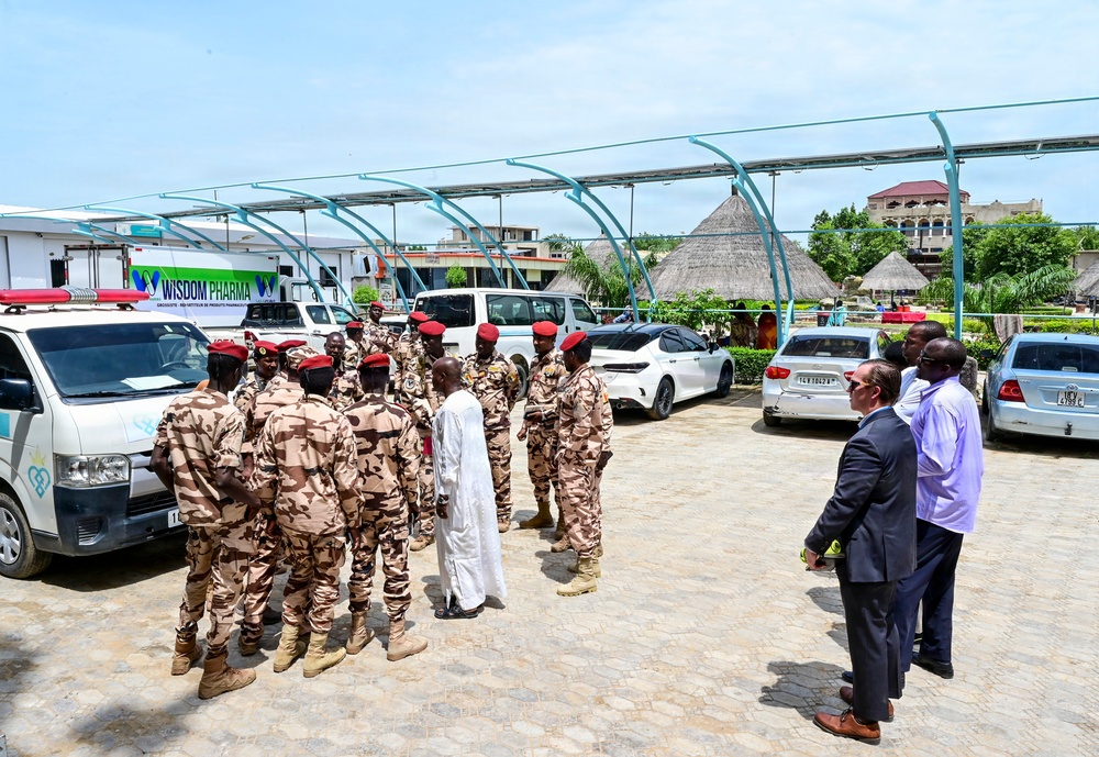 A group of military personnel in desert camouflage uniforms and red berets are gathered in a paved outdoor area alongside a civilian in white traditional attire. Two men in civilian dress — one in a dark suit and one in a light purple shirt — stand separately to the right observing the group. The setting includes parked vehicles, a solar panel canopy structure, and a banner reading 'Wisdom Pharma' in the background.