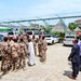 A group of military personnel in desert camouflage uniforms and red berets are gathered in a paved outdoor area alongside a civilian in white traditional attire. Two men in civilian dress — one in a dark suit and one in a light purple shirt — stand separately to the right observing the group. The setting includes parked vehicles, a solar panel canopy structure, and a banner reading 'Wisdom Pharma' in the background.
