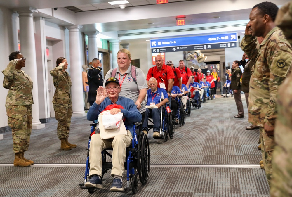 A group of elderly veterans in blue wheelchairs are being escorted through an airport terminal corridor. Active-duty military personnel in camouflage uniforms line both sides of the hallway, rendering salutes as the veterans pass. This appears to be a Honor Flight event, where veterans are honored and transported to visit war memorials.