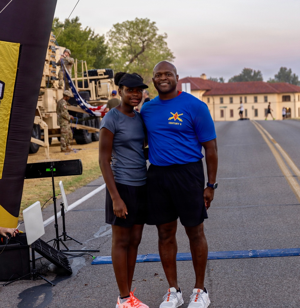 Two individuals pose together near what appears to be a race start/finish line on a military installation. A man wearing a blue 'Century 6' t-shirt with military insignia and a woman in a gray t-shirt and visor stand together smiling. In the background, military vehicles, soldiers, and an American flag are visible alongside institutional brick buildings.