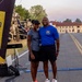 Two individuals pose together near what appears to be a race start/finish line on a military installation. A man wearing a blue 'Century 6' t-shirt with military insignia and a woman in a gray t-shirt and visor stand together smiling. In the background, military vehicles, soldiers, and an American flag are visible alongside institutional brick buildings.