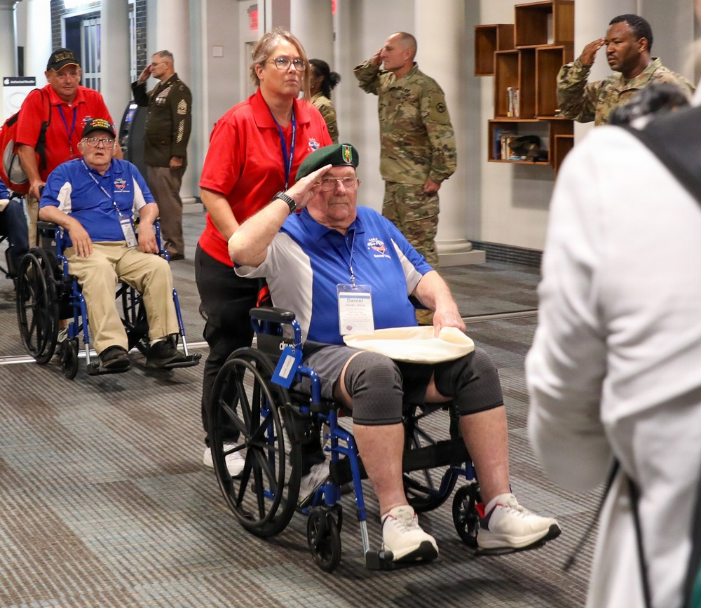 A group of elderly veterans in wheelchairs are being escorted through what appears to be a military or veterans' facility, likely during an honor flight or similar tribute event. A veteran in the foreground wearing a green beret and blue polo shirt is rendering a salute while being pushed by a volunteer in a red polo shirt. Active-duty military personnel in camouflage uniforms are saluting the veterans in return.
