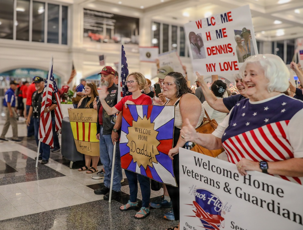 A crowd of people gathered in what appears to be an airport terminal to welcome home veterans and their guardians as part of an Honor Flight event. Participants are holding handmade signs, American flags, and wearing patriotic clothing to greet returning veterans. The atmosphere appears celebratory and emotionally charged, with visible smiles and enthusiastic gestures.