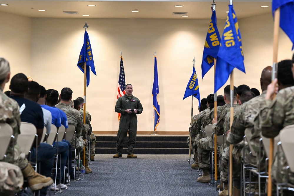 A military officer in olive/green flight suit stands at the center of an aisle addressing seated personnel during a formal military ceremony or briefing. The setting appears to be an indoor auditorium or conference hall with multiple blue unit guidon flags flanking the aisle. The flags visible include markings consistent with the 403rd Wing (403 WG) and related units.