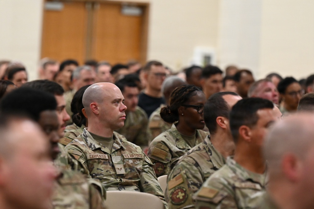 A large group of U.S. Air Force personnel in OCP (Operational Camouflage Pattern) uniforms are seated in an auditorium or briefing hall, appearing to attend a formal presentation or briefing. The central figure is identified by a name tape as 'Carpenter' and wears U.S. Air Force insignia. The crowd is diverse and attentive, suggesting an all-hands meeting or official military event.