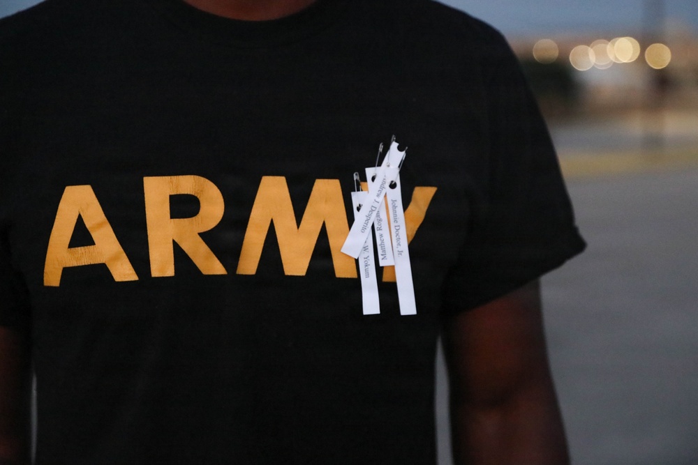 A person wearing a black Army t-shirt with gold lettering has several white paper strips pinned to the chest area with safety pins. The strips appear to contain printed names, suggesting a memorial or commemorative gesture. The background is blurred with bokeh lights, indicating an outdoor evening setting.