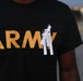 A person wearing a black Army t-shirt with gold lettering has several white paper strips pinned to the chest area with safety pins. The strips appear to contain printed names, suggesting a memorial or commemorative gesture. The background is blurred with bokeh lights, indicating an outdoor evening setting.