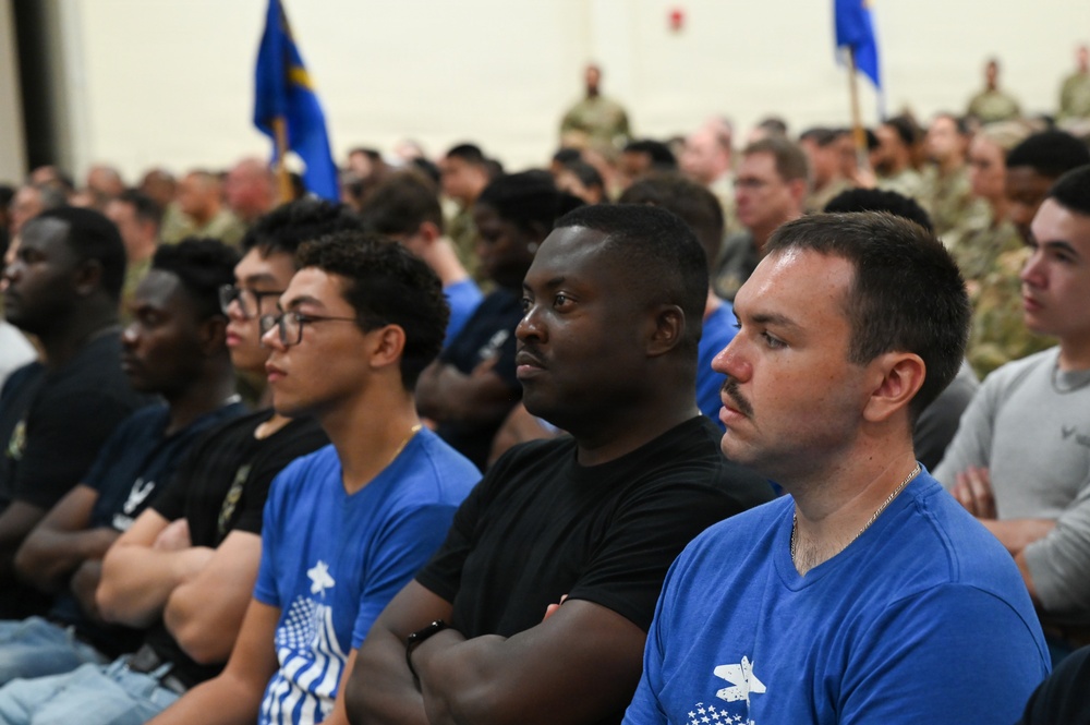 A large group of military personnel and civilians are seated in an assembly hall or hangar, attentively listening to a presentation or briefing. The foreground shows individuals wearing blue t-shirts with what appears to be a U.S. Air Force insignia, while uniformed personnel in camouflage are visible in the background. The setting suggests a formal military gathering, orientation, or all-hands meeting.