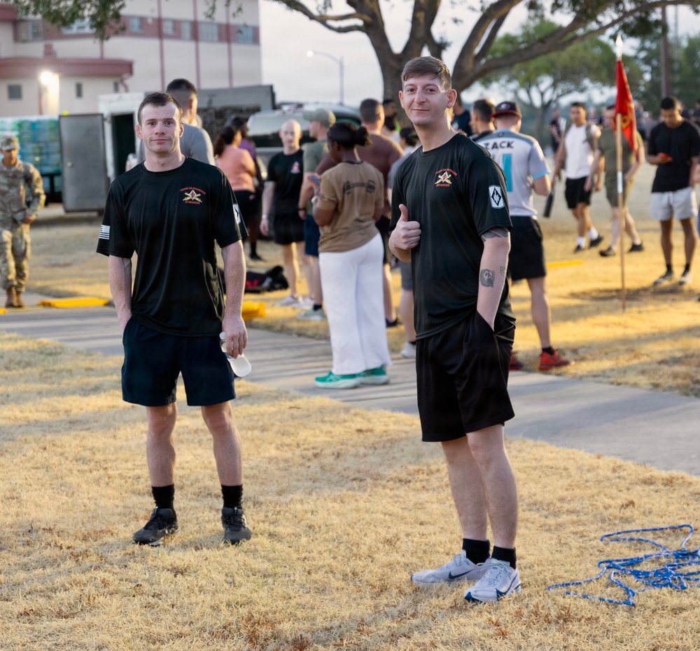 Two young men in matching black military physical training (PT) uniforms stand in the foreground on a dry grass area, one giving a thumbs-up to the camera. Behind them, a large group of people in various athletic and military attire are gathered on what appears to be a military installation or base. A red military guidon flag is visible in the background along with institutional buildings.