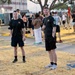 Two young men in matching black military physical training (PT) uniforms stand in the foreground on a dry grass area, one giving a thumbs-up to the camera. Behind them, a large group of people in various athletic and military attire are gathered on what appears to be a military installation or base. A red military guidon flag is visible in the background along with institutional buildings.