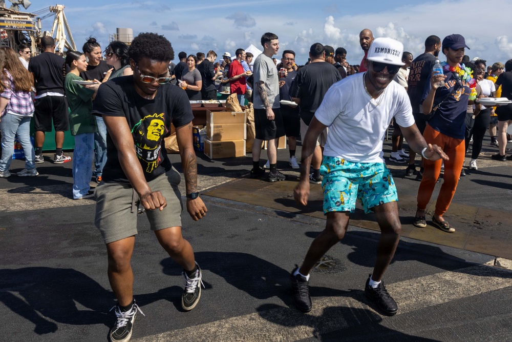 Two men are dancing energetically in the foreground at what appears to be an outdoor social gathering or deck party. One wears a Bob Marley graphic t-shirt with khaki shorts, while the other wears a white t-shirt, teal tropical-print shorts, and a white cap. A large crowd of people mingles in the background near food/beverage tables on what appears to be a ship's deck.