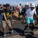 Two men are dancing energetically in the foreground at what appears to be an outdoor social gathering or deck party. One wears a Bob Marley graphic t-shirt with khaki shorts, while the other wears a white t-shirt, teal tropical-print shorts, and a white cap. A large crowd of people mingles in the background near food/beverage tables on what appears to be a ship's deck.