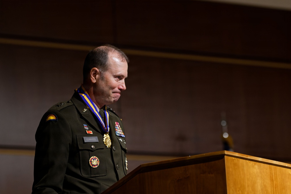 A U.S. Army officer in dress green uniform stands at a wooden podium, appearing visibly emotional. He is wearing a large medallion on a blue and yellow ribbon around his neck, along with multiple service ribbons and badges on his uniform. The setting appears to be a formal military ceremony, likely a retirement or awards ceremony.