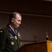 A U.S. Army officer in dress green uniform stands at a wooden podium, appearing visibly emotional. He is wearing a large medallion on a blue and yellow ribbon around his neck, along with multiple service ribbons and badges on his uniform. The setting appears to be a formal military ceremony, likely a retirement or awards ceremony.