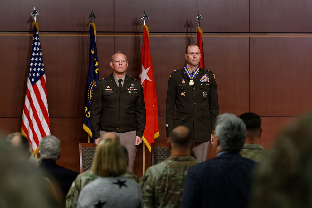 A formal U.S. Army military ceremony featuring two officers standing at attention on a stage before an audience. The officer on the right is wearing a large medallion/award around his neck, suggesting this is a decoration or retirement ceremony. Several flags including the U.S. flag, a state flag, and red general officer flags are displayed in the background.