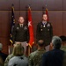 A formal U.S. Army military ceremony featuring two officers standing at attention on a stage before an audience. The officer on the right is wearing a large medallion/award around his neck, suggesting this is a decoration or retirement ceremony. Several flags including the U.S. flag, a state flag, and red general officer flags are displayed in the background.