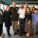 A group of approximately nine people pose together for a formal group photograph in what appears to be an indoor facility such as a library or museum. One individual is wearing a U.S. Army dress uniform with multiple service ribbons and medals, suggesting a military ceremony or recognition event. The group includes adults and at least one child, dressed in a mix of formal and semi-formal attire.