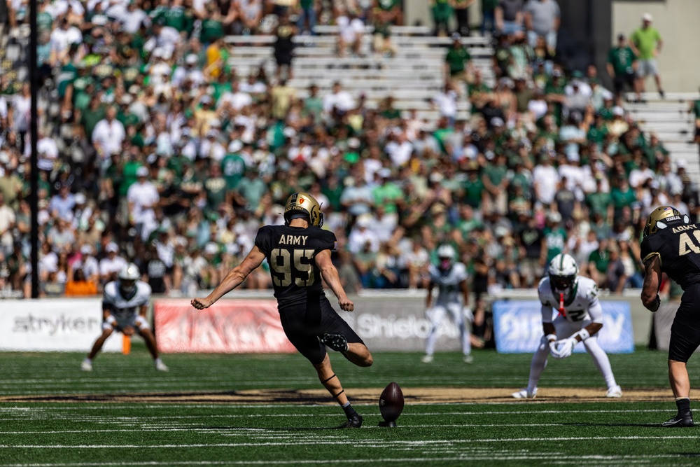 An Army West Point football player wearing jersey number 95 is captured mid-kick during a college football game, likely a kickoff or field goal attempt. The opposing team, dressed in white and green uniforms, is visible in the background along with a packed stadium of spectators. The game appears to be played at a well-attended outdoor stadium in daylight conditions.