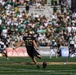 An Army West Point football player wearing jersey number 95 is captured mid-kick during a college football game, likely a kickoff or field goal attempt. The opposing team, dressed in white and green uniforms, is visible in the background along with a packed stadium of spectators. The game appears to be played at a well-attended outdoor stadium in daylight conditions.