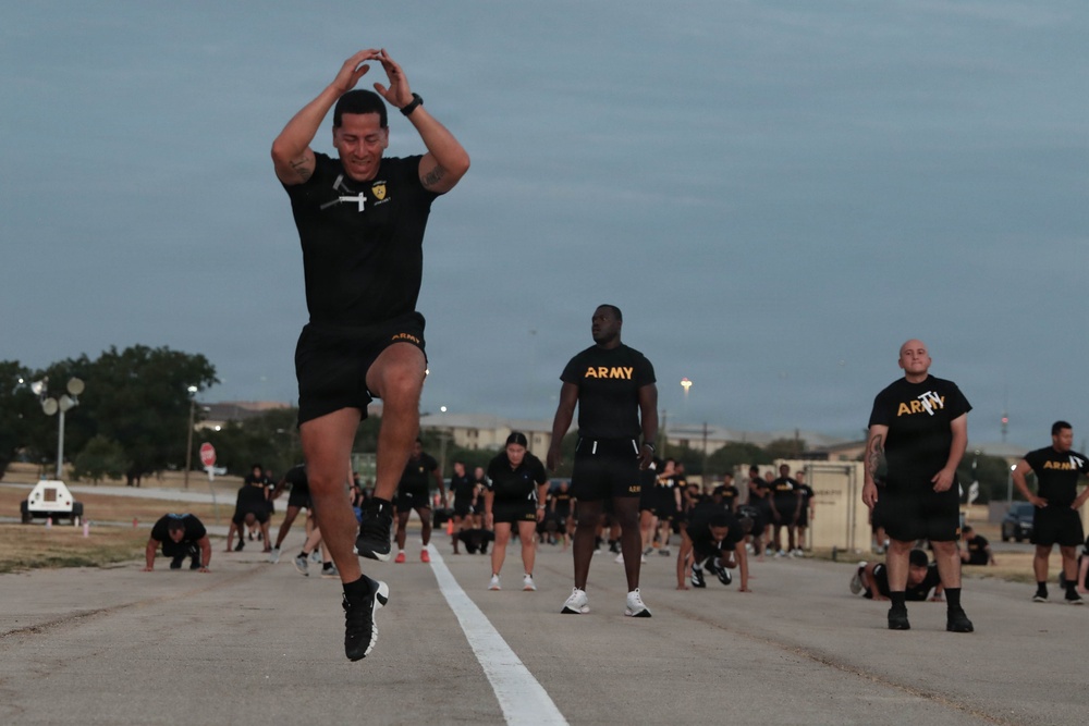A group of U.S. Army soldiers in black PT (physical training) uniforms are conducting an outdoor physical fitness exercise session on a paved road or runway, likely at a military installation. The foreground features a soldier mid-air performing what appears to be a jumping jack or similar calisthenic movement with hands raised overhead. The scene takes place during early morning or dusk conditions based on the ambient lighting.