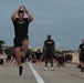 A group of U.S. Army soldiers in black PT (physical training) uniforms are conducting an outdoor physical fitness exercise session on a paved road or runway, likely at a military installation. The foreground features a soldier mid-air performing what appears to be a jumping jack or similar calisthenic movement with hands raised overhead. The scene takes place during early morning or dusk conditions based on the ambient lighting.