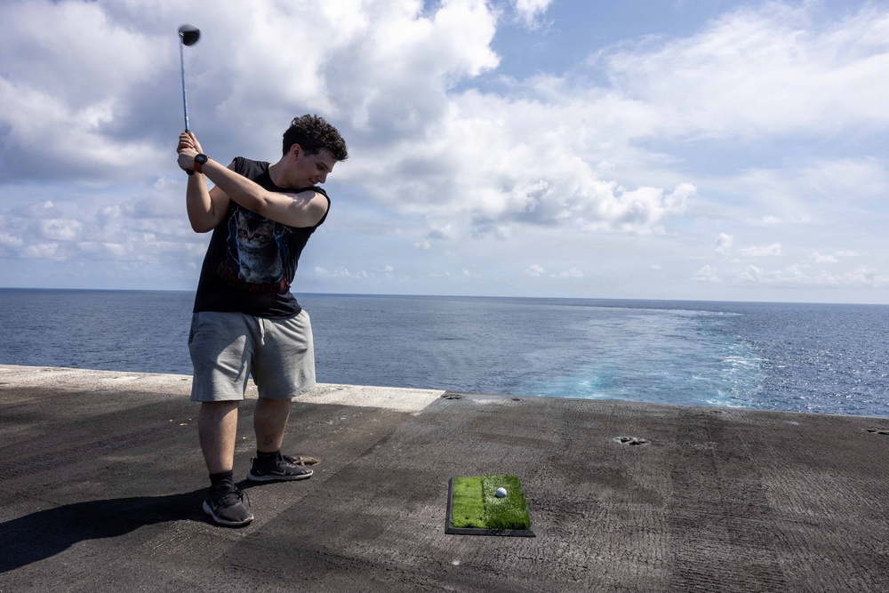 A young man is swinging a golf driver on what appears to be the deck of a naval vessel or large ship, with the open ocean visible in the background. He is using a small portable artificial turf mat with a golf ball placed on it. The setting creates an unusual juxtaposition of a recreational golf activity in a maritime environment.