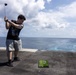 A young man is swinging a golf driver on what appears to be the deck of a naval vessel or large ship, with the open ocean visible in the background. He is using a small portable artificial turf mat with a golf ball placed on it. The setting creates an unusual juxtaposition of a recreational golf activity in a maritime environment.