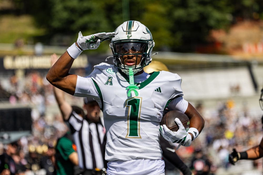 A college football player wearing jersey number 1 in white and green uniform performs a salute celebration while holding a football, likely after scoring a touchdown. The player appears to represent the University of South Florida (USF) Bulls, identifiable by the helmet logo and green color scheme. A referee in a black and white striped shirt is visible in the background along with a crowd of spectators.