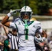 A college football player wearing jersey number 1 in white and green uniform performs a salute celebration while holding a football, likely after scoring a touchdown. The player appears to represent the University of South Florida (USF) Bulls, identifiable by the helmet logo and green color scheme. A referee in a black and white striped shirt is visible in the background along with a crowd of spectators.