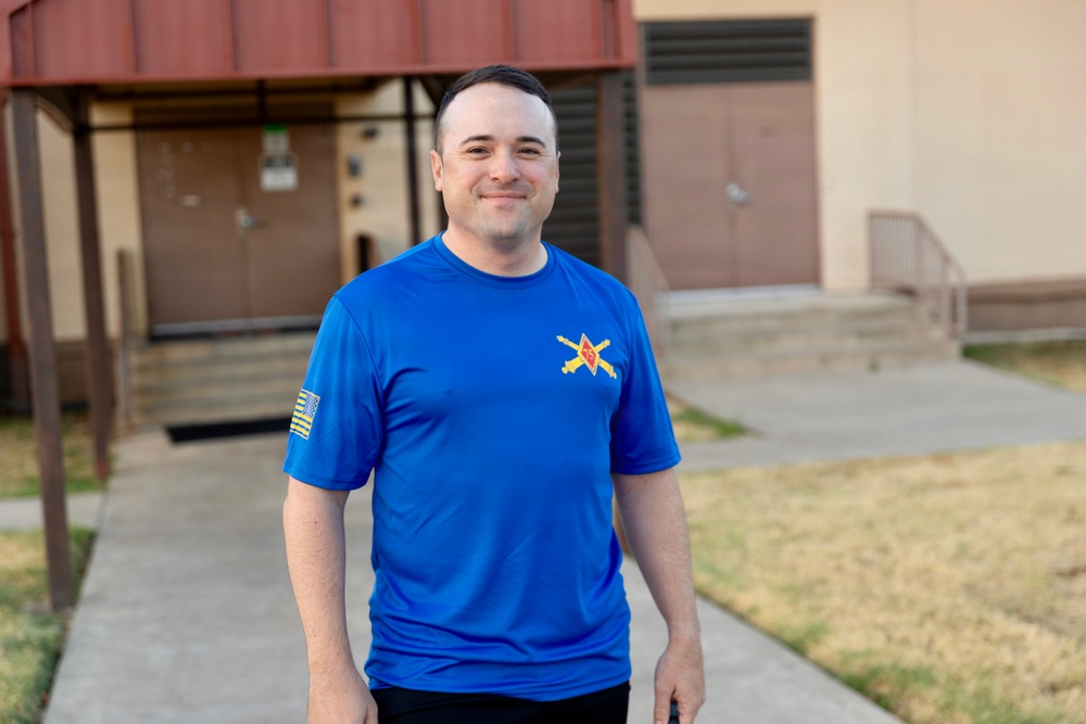 A man stands outdoors on a concrete walkway in front of a building, smiling at the camera. He is wearing a bright blue athletic t-shirt featuring a military-style crossed-sabers insignia on the chest and what appears to be a subdued American flag patch on the left sleeve. The setting appears to be a military installation or institutional campus.