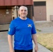 A man stands outdoors on a concrete walkway in front of a building, smiling at the camera. He is wearing a bright blue athletic t-shirt featuring a military-style crossed-sabers insignia on the chest and what appears to be a subdued American flag patch on the left sleeve. The setting appears to be a military installation or institutional campus.
