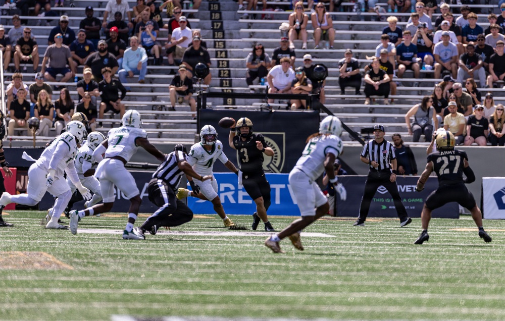 A college football game is in progress, showing a quarterback in a black uniform (number 3) releasing a pass while under pressure from defenders in white uniforms. The home team appears to be Army (West Point), identifiable by the black uniforms and 'ARMY' text visible on jersey number 27. The game is being played in a stadium with a moderately filled crowd watching under bright daylight conditions.