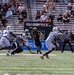 A college football game is in progress, showing a quarterback in a black uniform (number 3) releasing a pass while under pressure from defenders in white uniforms. The home team appears to be Army (West Point), identifiable by the black uniforms and 'ARMY' text visible on jersey number 27. The game is being played in a stadium with a moderately filled crowd watching under bright daylight conditions.