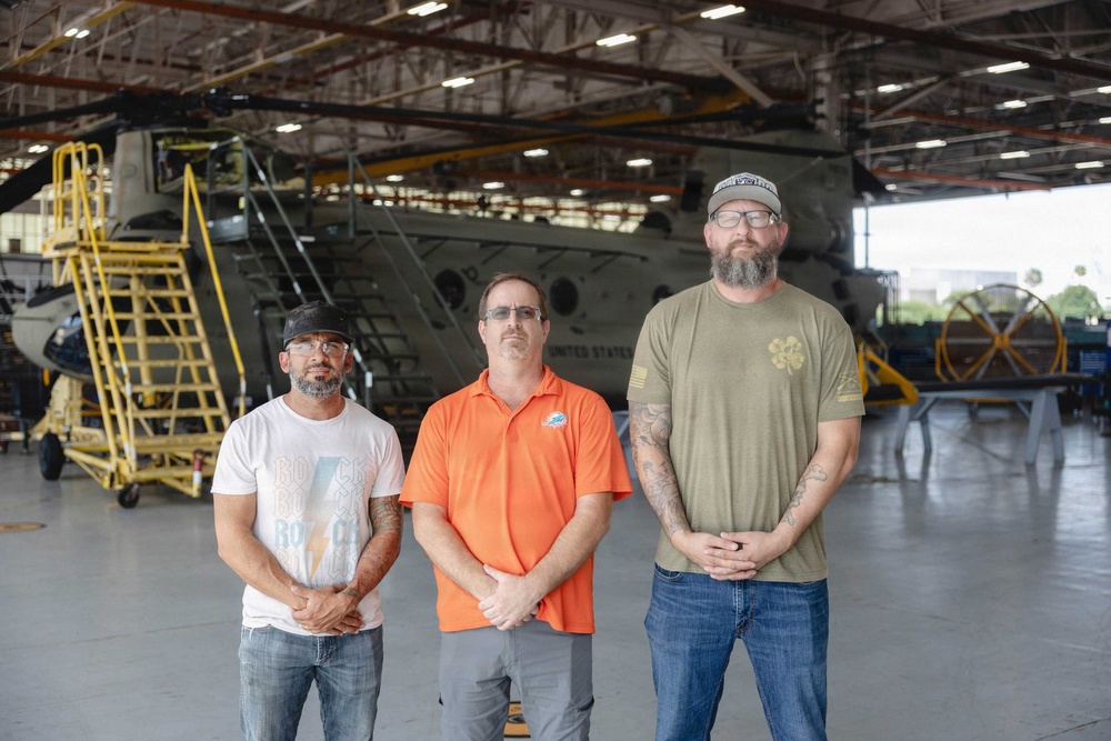 Three men stand posed together inside a large military aircraft hangar. Behind them is a large tandem-rotor helicopter, likely a CH-47 Chinook, marked with 'UNITED STATES' text. The setting appears to be a maintenance or storage facility based on the equipment and scaffolding visible.