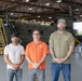 Three men stand posed together inside a large military aircraft hangar. Behind them is a large tandem-rotor helicopter, likely a CH-47 Chinook, marked with 'UNITED STATES' text. The setting appears to be a maintenance or storage facility based on the equipment and scaffolding visible.