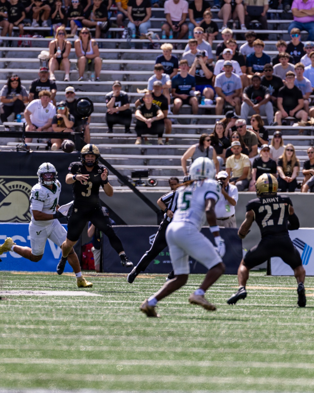 A college football game is in progress, showing an Army Black Knights quarterback wearing number 3 releasing a pass while under pressure from a defender wearing number 4 in white and green uniform. The ball is visible in mid-air above the players. Army players in black uniforms and opposing players in white/green uniforms are visible on the field, with a stadium crowd watching in the background.