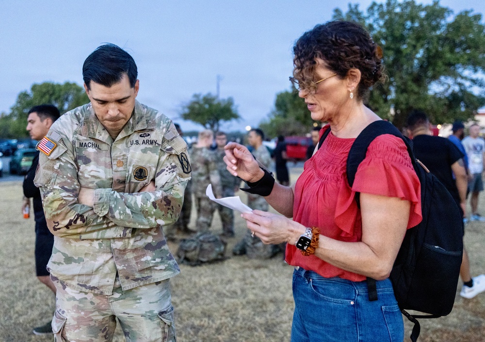 A U.S. Army soldier named Machal, wearing OCP (Operational Camouflage Pattern) uniform, stands with arms crossed while a civilian woman in a red top reviews a document. The scene appears to take place on a military installation during early morning or evening hours, with other soldiers and civilians visible in the background. The interaction suggests an administrative or informational exchange.