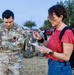 A U.S. Army soldier named Machal, wearing OCP (Operational Camouflage Pattern) uniform, stands with arms crossed while a civilian woman in a red top reviews a document. The scene appears to take place on a military installation during early morning or evening hours, with other soldiers and civilians visible in the background. The interaction suggests an administrative or informational exchange.