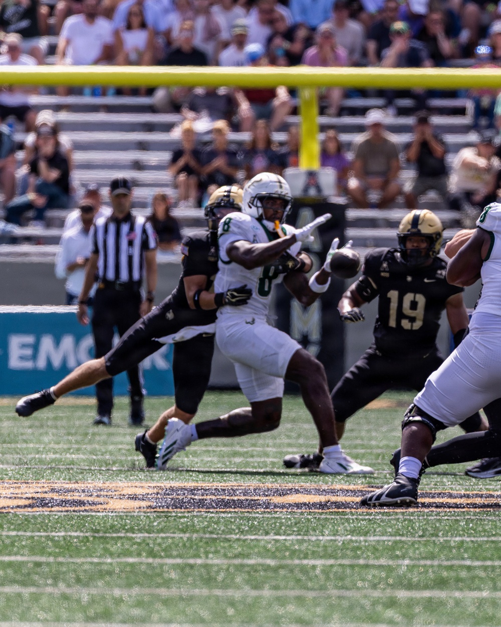 A college football game in progress, showing a player in a white uniform (number 8) appearing to throw or lateral a pass while being pressured by defenders in black uniforms. Players wearing black jerseys with numbers 8 and 19 are visible on defense, along with a referee in a striped shirt observing the play. The action takes place near the end zone, indicated by the visible yellow goal post in the background.