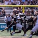 A college football game in progress, showing a player in a white uniform (number 8) appearing to throw or lateral a pass while being pressured by defenders in black uniforms. Players wearing black jerseys with numbers 8 and 19 are visible on defense, along with a referee in a striped shirt observing the play. The action takes place near the end zone, indicated by the visible yellow goal post in the background.