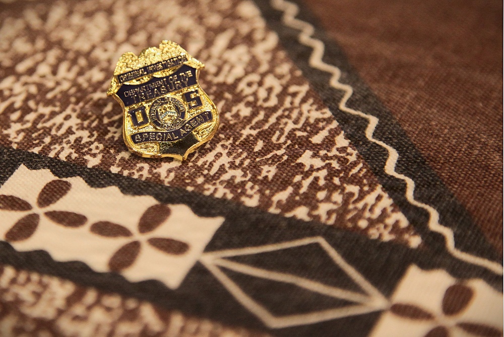 A small gold and blue law enforcement lapel pin rests on a patterned brown and cream fabric, likely a batik or woven textile. The pin bears the insignia of the U.S. Department of the Treasury, Criminal Investigation division, identifying the holder as a Special Agent. The image is a close-up shot with shallow depth of field, emphasizing the badge against the decorative textile background.