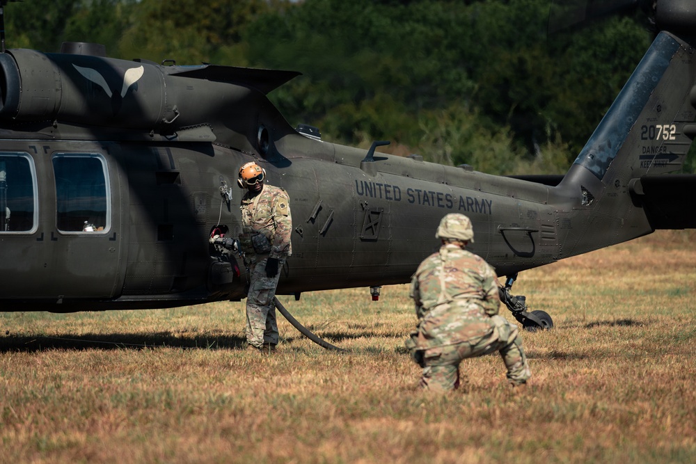 Two U.S. Army soldiers are conducting a forward area refueling point (FARP) operation on a UH-60 Black Hawk helicopter in an open field. The helicopter bears the tail number 20752 and 'UNITED STATES ARMY' markings on its fuselage. One soldier wearing an orange flight helmet is managing a fuel hose near the aircraft while a second soldier in standard combat gear approaches.