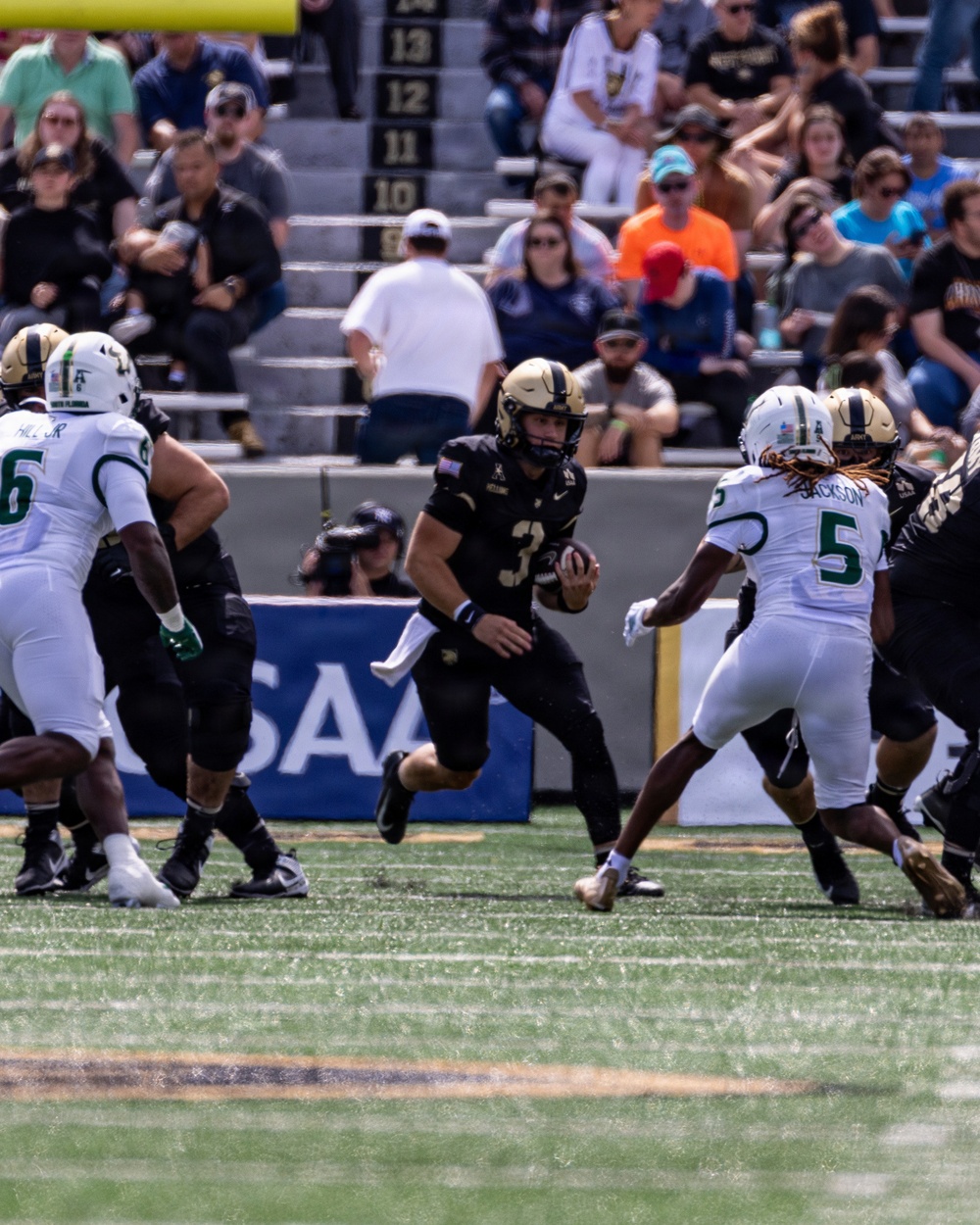 A college football game in progress showing a ball carrier in a black uniform (number 3) running through a gap between defenders. The offensive team wears black and gold uniforms consistent with Army West Point, while the defensive team wears white and green uniforms with visible name 'JACKSON' and number 5. The game is being played in a stadium with spectators visible in the stands.