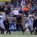 A college football game in progress showing a ball carrier in a black uniform (number 3) running through a gap between defenders. The offensive team wears black and gold uniforms consistent with Army West Point, while the defensive team wears white and green uniforms with visible name 'JACKSON' and number 5. The game is being played in a stadium with spectators visible in the stands.
