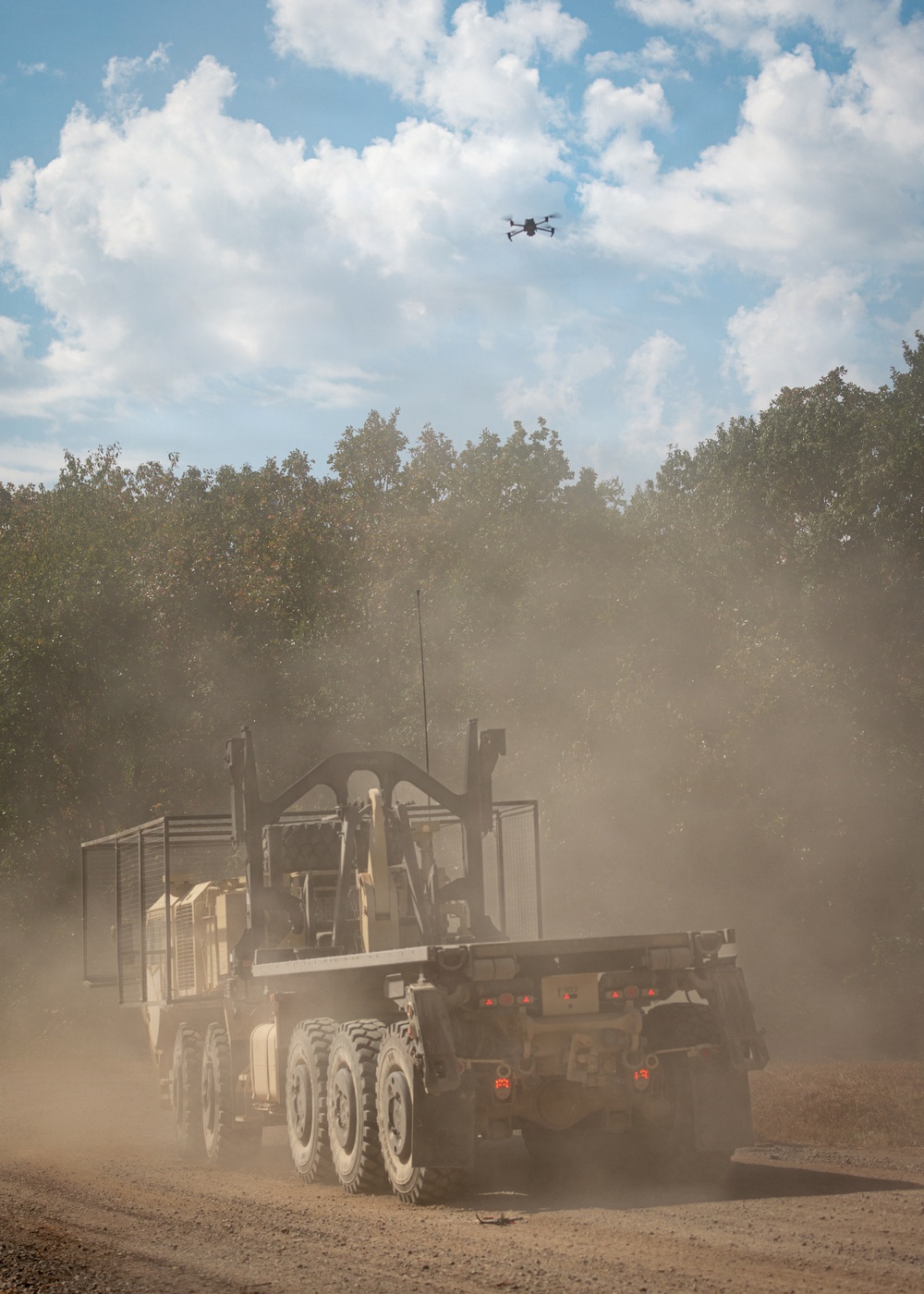 A large military heavy-duty truck, viewed from the rear, travels along a dusty dirt road kicking up a significant dust cloud. A quadcopter drone is visible hovering in the sky above the scene. The setting appears to be a training area or field exercise environment bordered by trees under a partly cloudy sky.