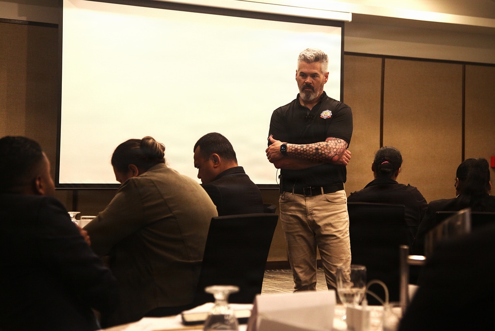 A man with gray hair and a beard stands at the front of a conference or training room with arms crossed, appearing to lead or instruct a group of seated attendees. He is wearing a black polo shirt with a logo patch and khaki pants, with a visible geometric tattoo sleeve on his right arm. A large projection screen is visible behind him, currently displaying a mostly blank/white slide.