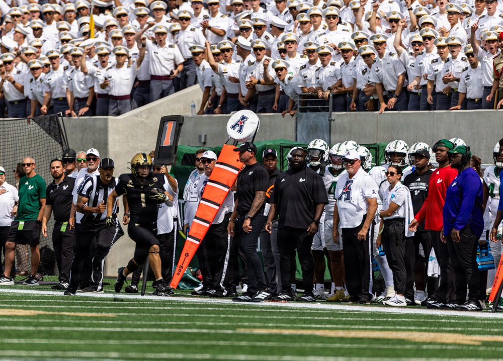 A college football game is in progress on a green turf field, featuring a sideline scene with players, coaches, and officials. One team wears black uniforms while the other wears white and green uniforms with an 'A' logo visible on helmets, consistent with the American Athletic Conference (AAC). In the background, a large section of uniformed cadets in white dress uniforms fills the stands, suggesting this may be an Army (West Point) home game.