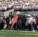 A college football game is in progress on a green turf field, featuring a sideline scene with players, coaches, and officials. One team wears black uniforms while the other wears white and green uniforms with an 'A' logo visible on helmets, consistent with the American Athletic Conference (AAC). In the background, a large section of uniformed cadets in white dress uniforms fills the stands, suggesting this may be an Army (West Point) home game.