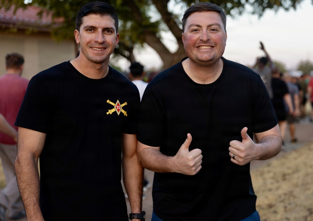 Two men in black t-shirts pose together outdoors, smiling at the camera. The man on the right is giving two thumbs up and wearing a ring. The left man's shirt displays a military-style insignia with crossed cannons and the number 75, suggesting an artillery unit affiliation.